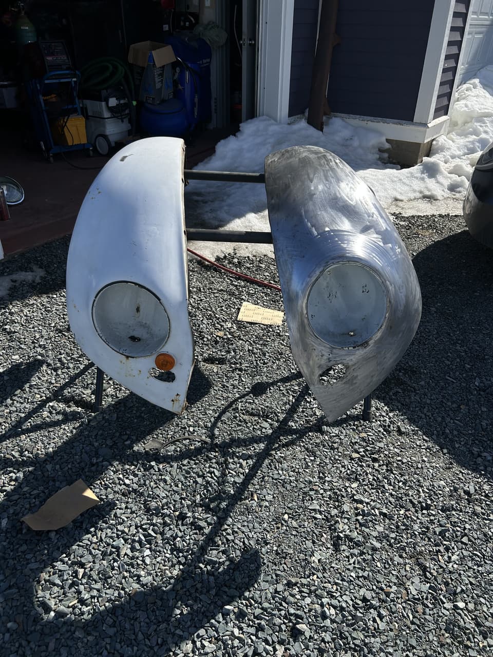 Two Beetle front fenders standing on gravel, the left one in old white paint and the right one stripped to bare metal
