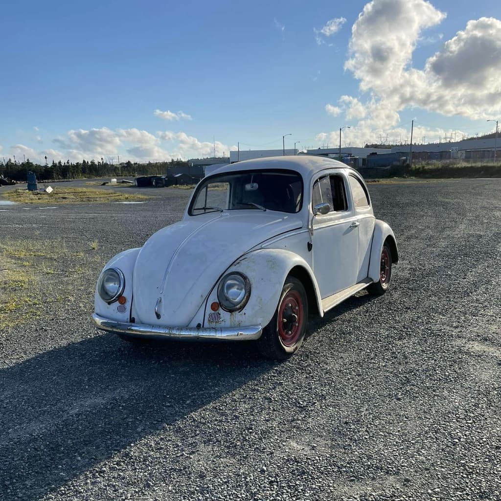 White 1964 Volkswagen Beetle with red wheels parked on gravel under a blue sky