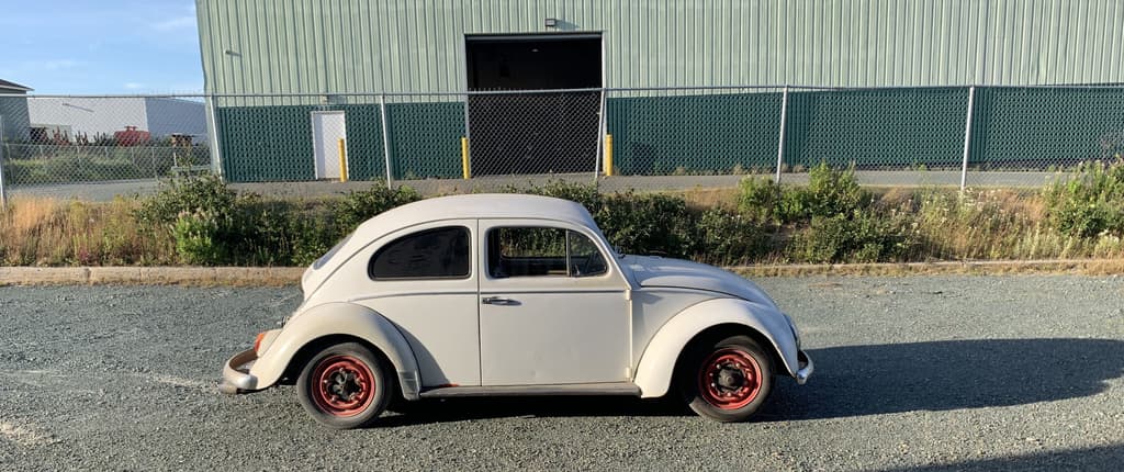 Side profile of the white 1964 Beetle with red wheels, parked on gravel in front of a green industrial building