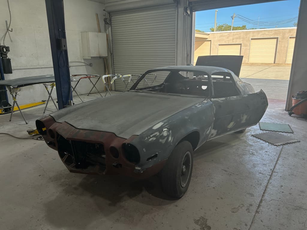 Front three-quarter view of the Camaro in grey high-build primer with lighter sanded patches, parked in the open shop doorway