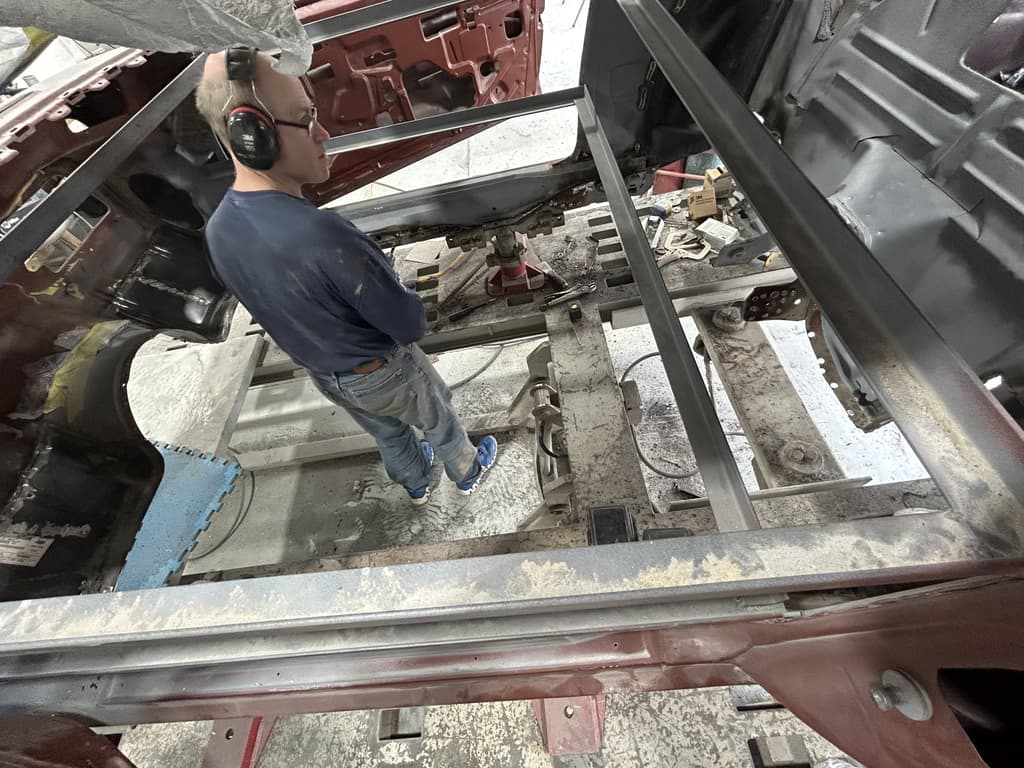 Camaro body braced with square steel tube on a frame bench, floors cut out, with a man in ear defenders standing in the opening