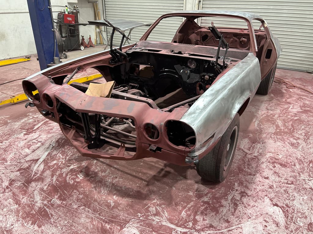 Front three-quarter view of the bare-metal Camaro body with the engine bay empty and the red oxide nose section still attached