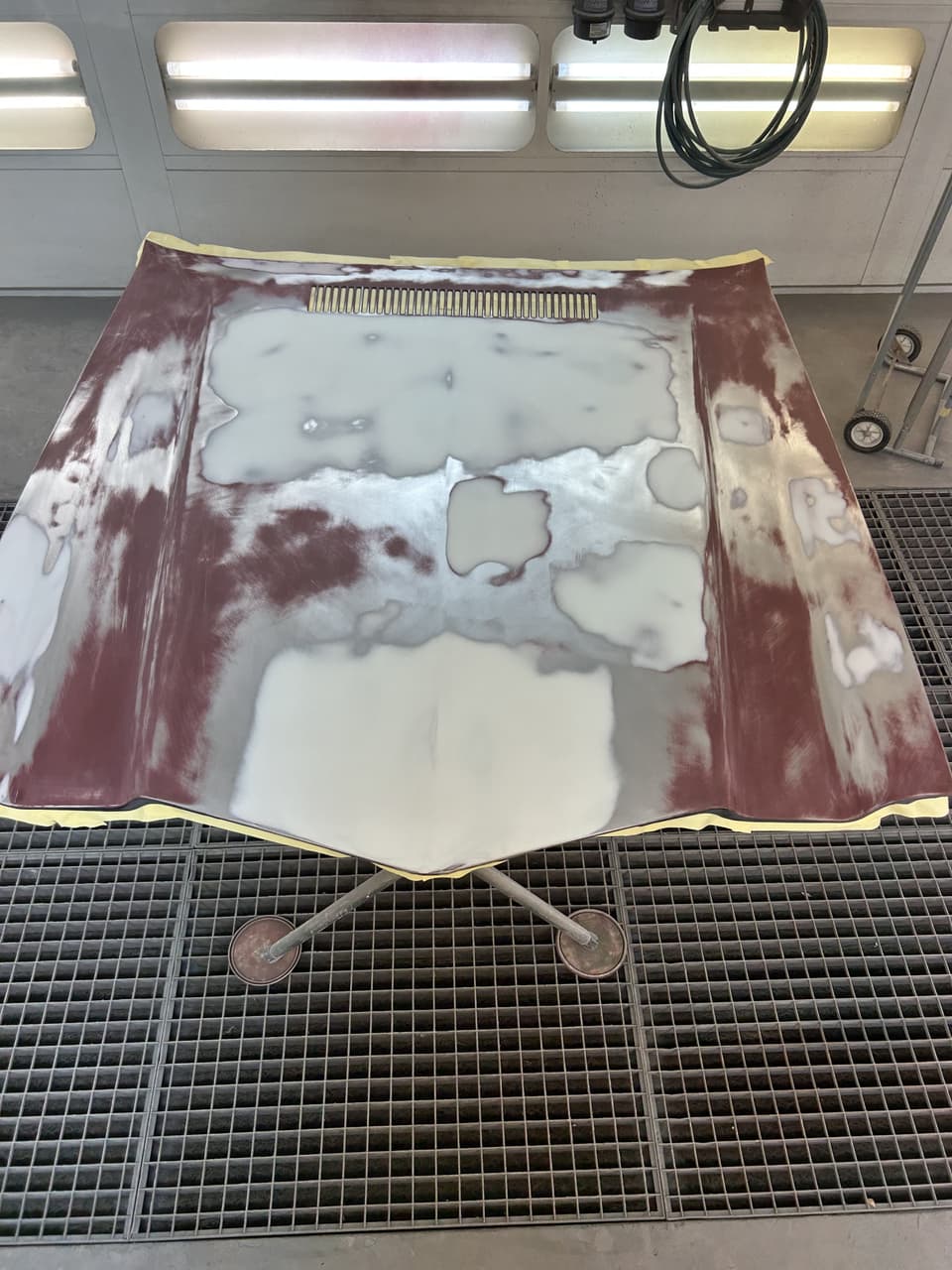 Flat Camaro hood on a stand in the spray booth, patches of red primer and grey filler partly sanded away to bare steel