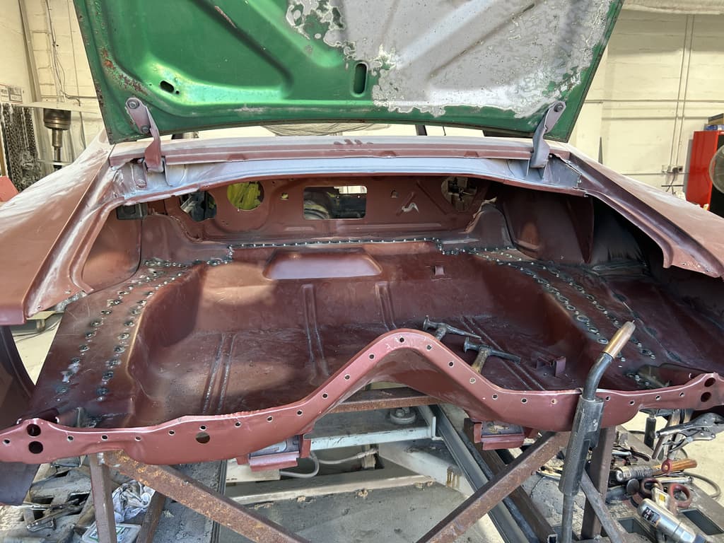 Trunk floor and rear frame rail in red primer with rows of weld spots along the seams and a welding torch tip in the corner of the frame
