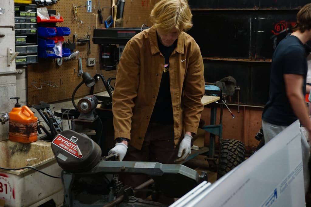 Patrick in a work jacket and gloves feeding steel tube into a horizontal bandsaw in the team shop