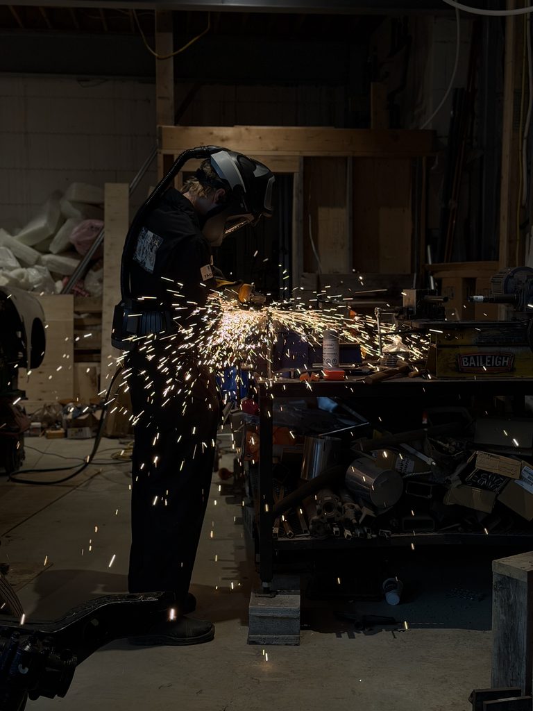 Patrick at a shop bench running an angle grinder on a tube held in a vise, throwing a spray of sparks