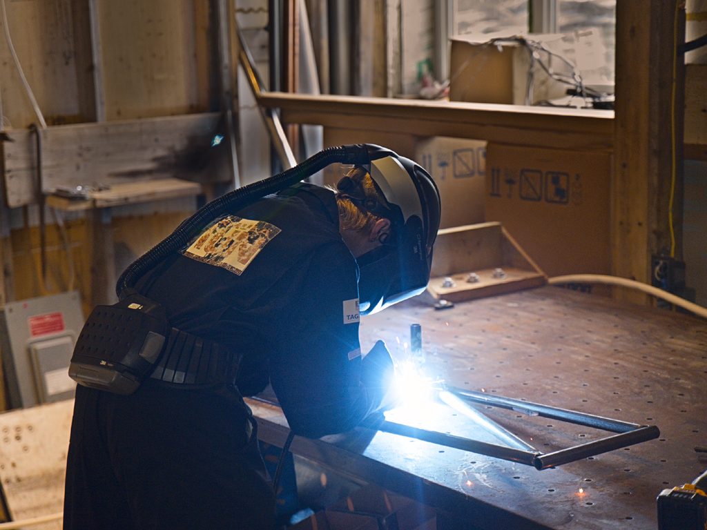 Patrick in a powered-air welding helmet welding steel tubes on a fixture table, with the arc lighting the joint