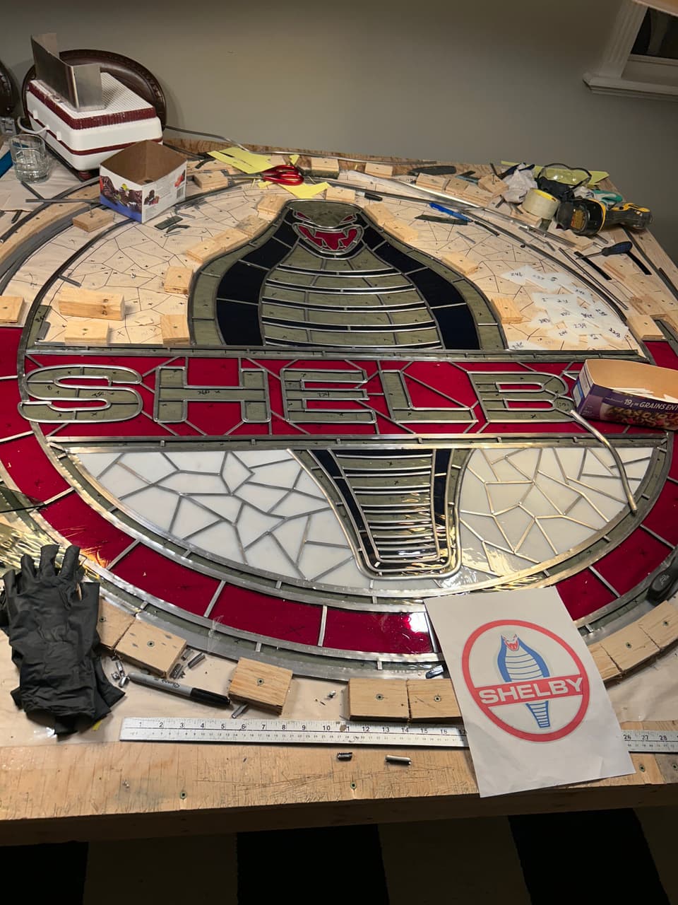 The Shelby panel during assembly on the workbench, held in by wooden strips, with a printed Shelby logo and a steel rule beside it